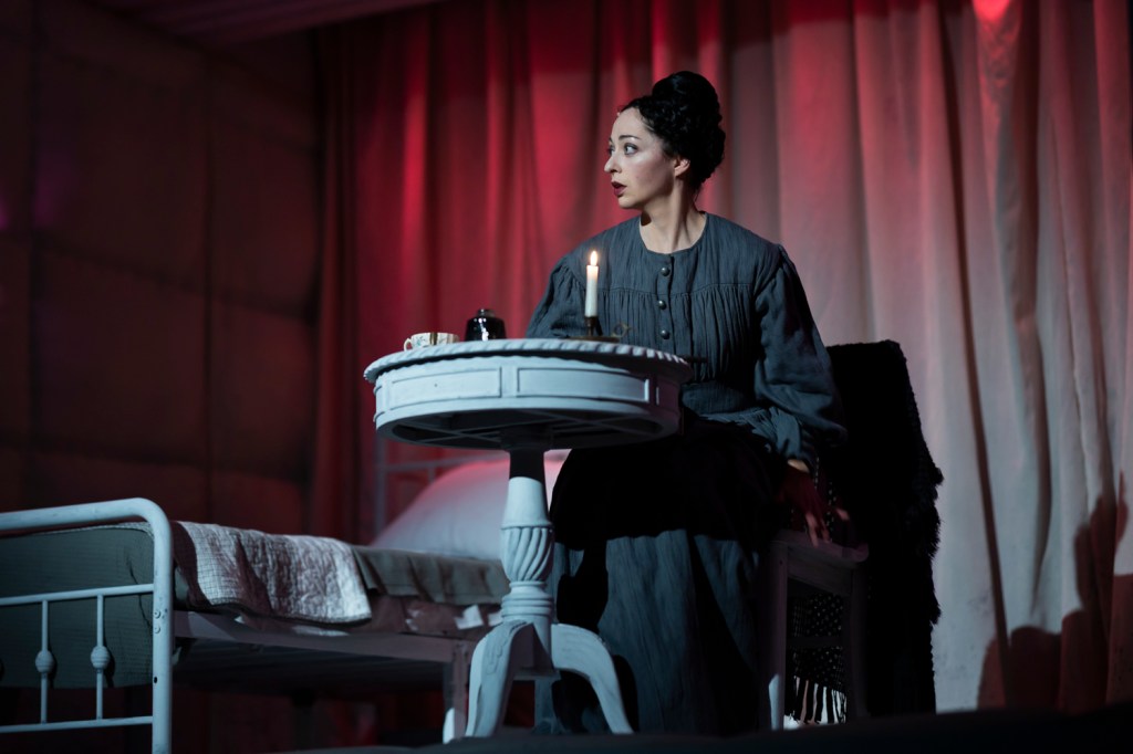 A woman in period clothing sits at a small table with a candle, looking to the side with a concerned expression, in a dimly lit room.