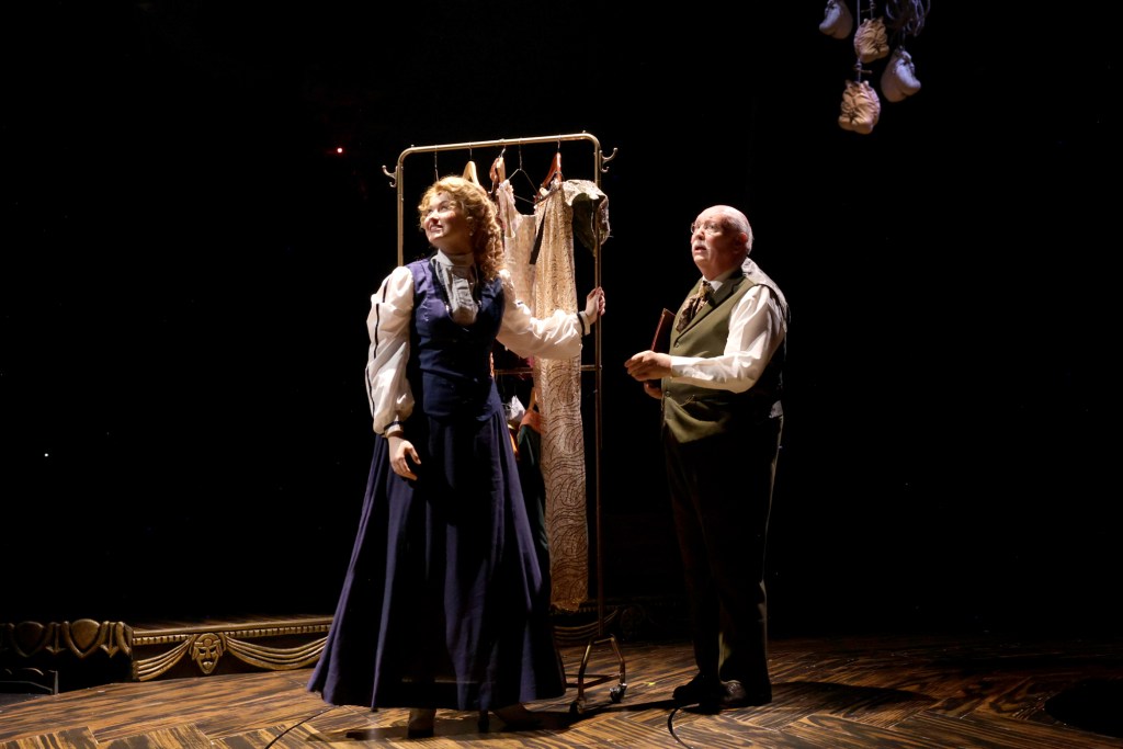 A scene from a theatrical performance featuring a woman in historical attire interacting with a man in formal clothing, with a garment rack in the background.