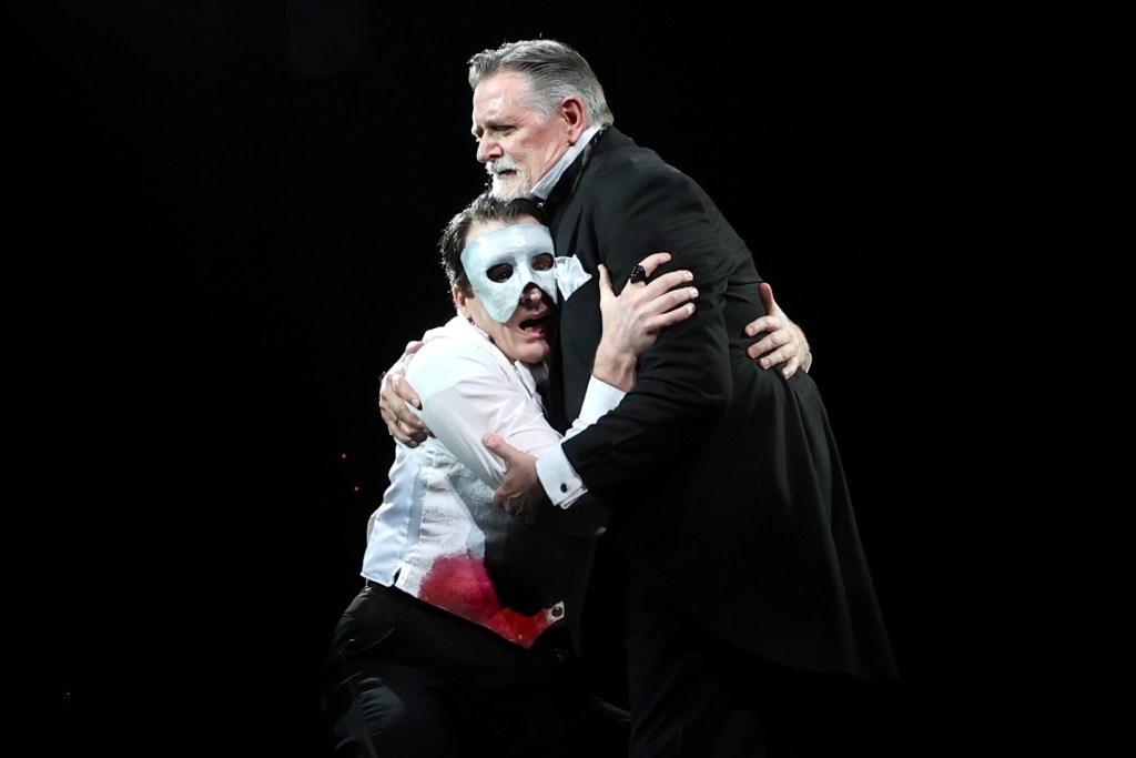 A dramatic moment between two male performers on stage, one dressed in formal wear with a black suit and the other in a white shirt with theatrical makeup and a white mask, embracing in an intense scene.