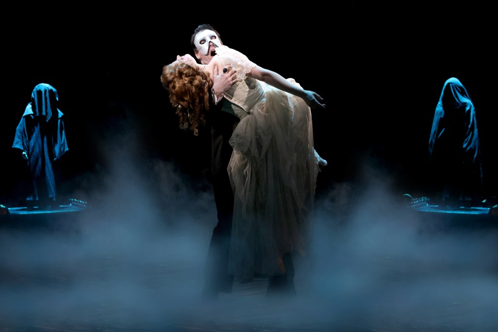 A theatrical scene featuring a man in a mask holding a woman in a flowing dress, surrounded by mist and shadowy figures in the background.